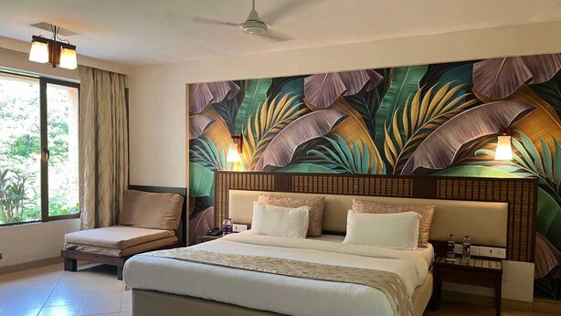 A hotel room with a bed, a chair, a nightstand, a ceiling fan, and a large window with curtains. - Silverador Resort Club (Mira Bhayandar)