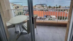 Varanda com mesa e cadeiras, vista para uma praia com edifícios e céu claro.