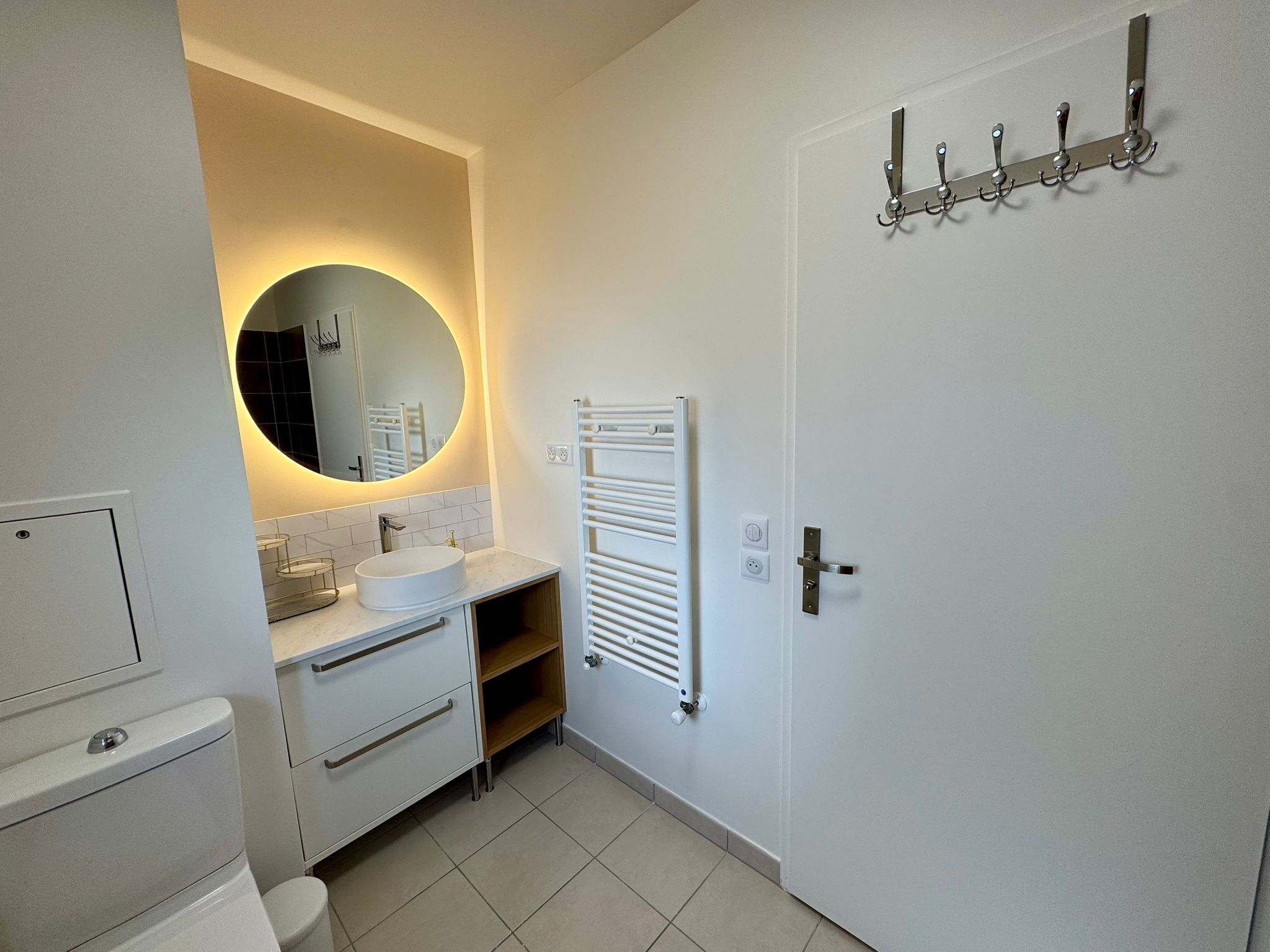 Een moderne badkamer met een ronde spiegel, een witte wastafel, een handdoekhouder en een deur met een handgreep.
