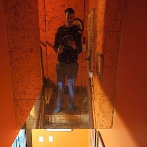 Une personne se tient devant un miroir, qui reflète un escalier, un mur à motifs et une porte.