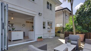 Een open terras met buitenmeubilair, een witte tafel en uitzicht op het interieur van het huis.