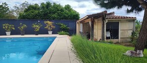 Une piscine dotée d’une terrasse carrelée, entourée de plantes en pot et bordée d’un petit bâtiment dont le toit est également recouvert de tuiles.