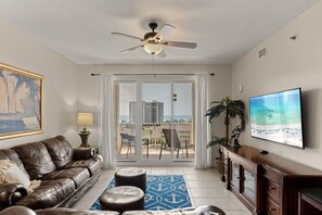 Ein Wohnzimmer mit einer Couch, einem Couchtisch, einem Fernseher, einem Deckenventilator und einem Balkon mit Blick auf Gebäude und Palmen.