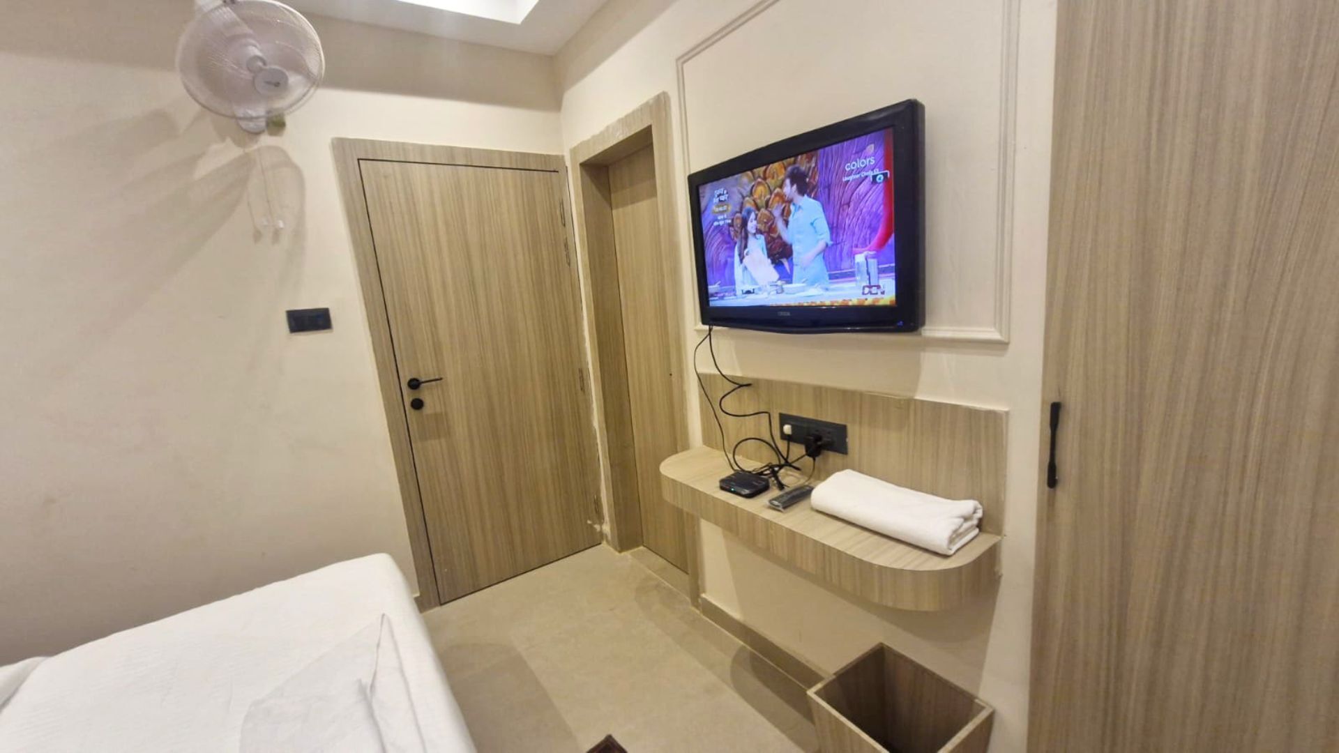 Une chambre d’hôtel avec un lit, un téléviseur fixé au mur, une petite étagère avec une télécommande et une porte.