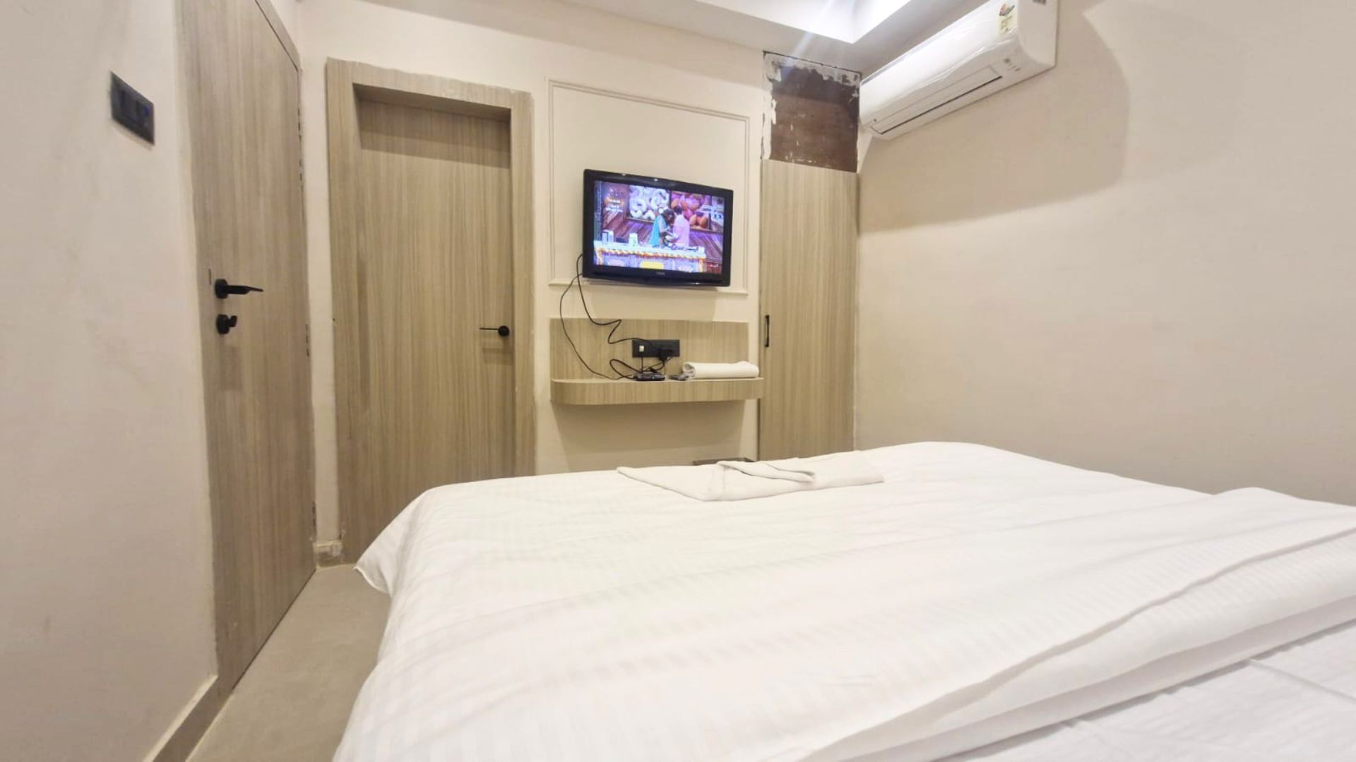 A hotel room with a bed, a television mounted on the wall, a door, and an air conditioning unit.
