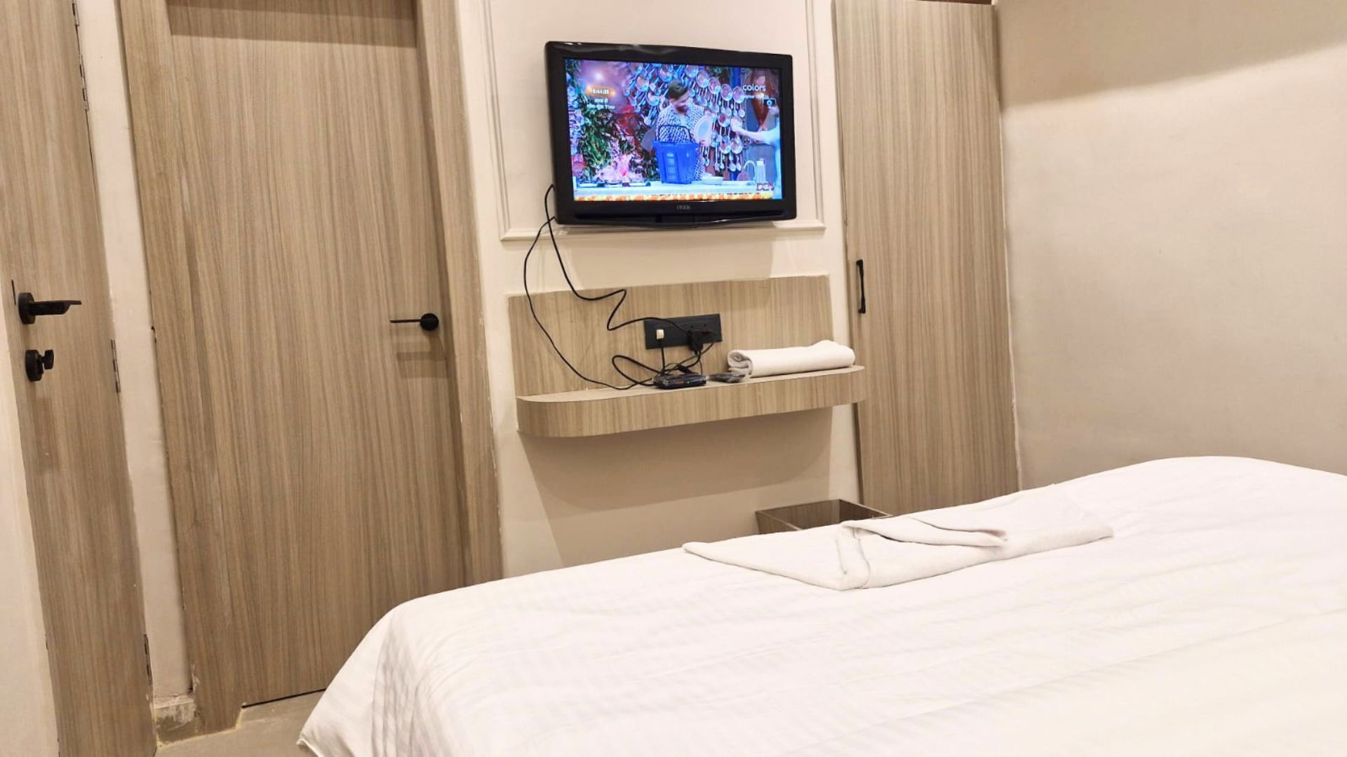 Une chambre d’hôtel avec un lit, un téléviseur fixé au mur, une étagère avec une serviette et une porte.