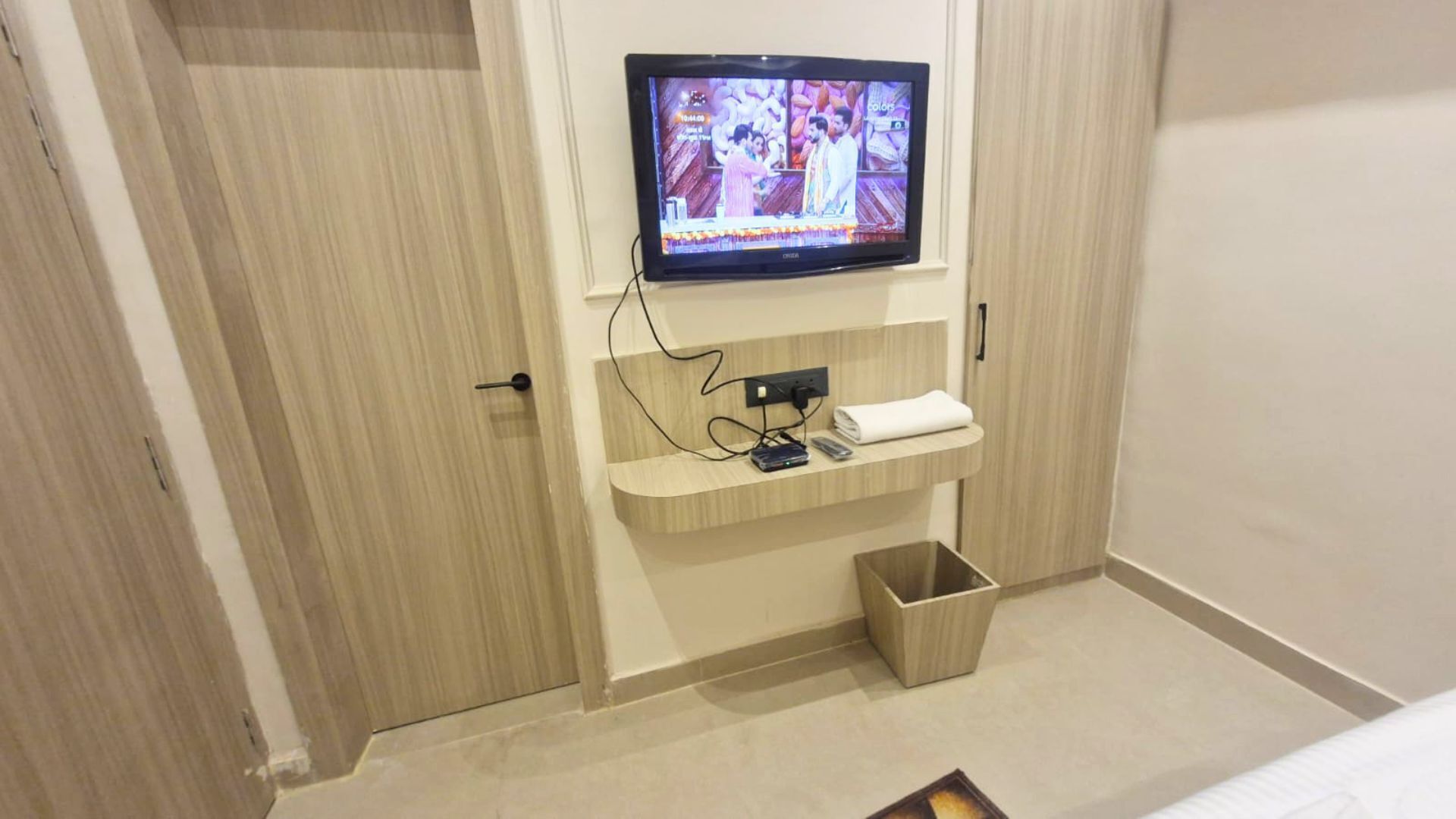 Quarto de hotel com televisão na parede, uma prateleira de madeira com uma toalha e um cesto de lixo.