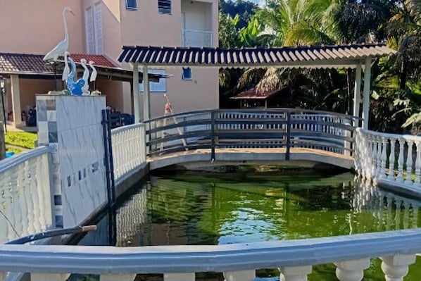 A bridge over a pond with a decorative sculpture, surrounded by a building with a tiled roof and a covered area. - Pousada Chácara Santa Inês (Conceição do Mato Dentro)