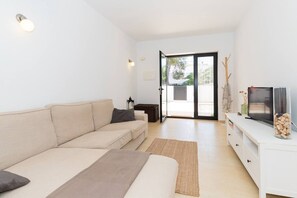 Ein modernes Wohnzimmer mit einer Couch, einem Flachbildfernseher, einem kleinen Tisch mit Dekoration und einer Glastür, die zu einem Außenbereich führt.