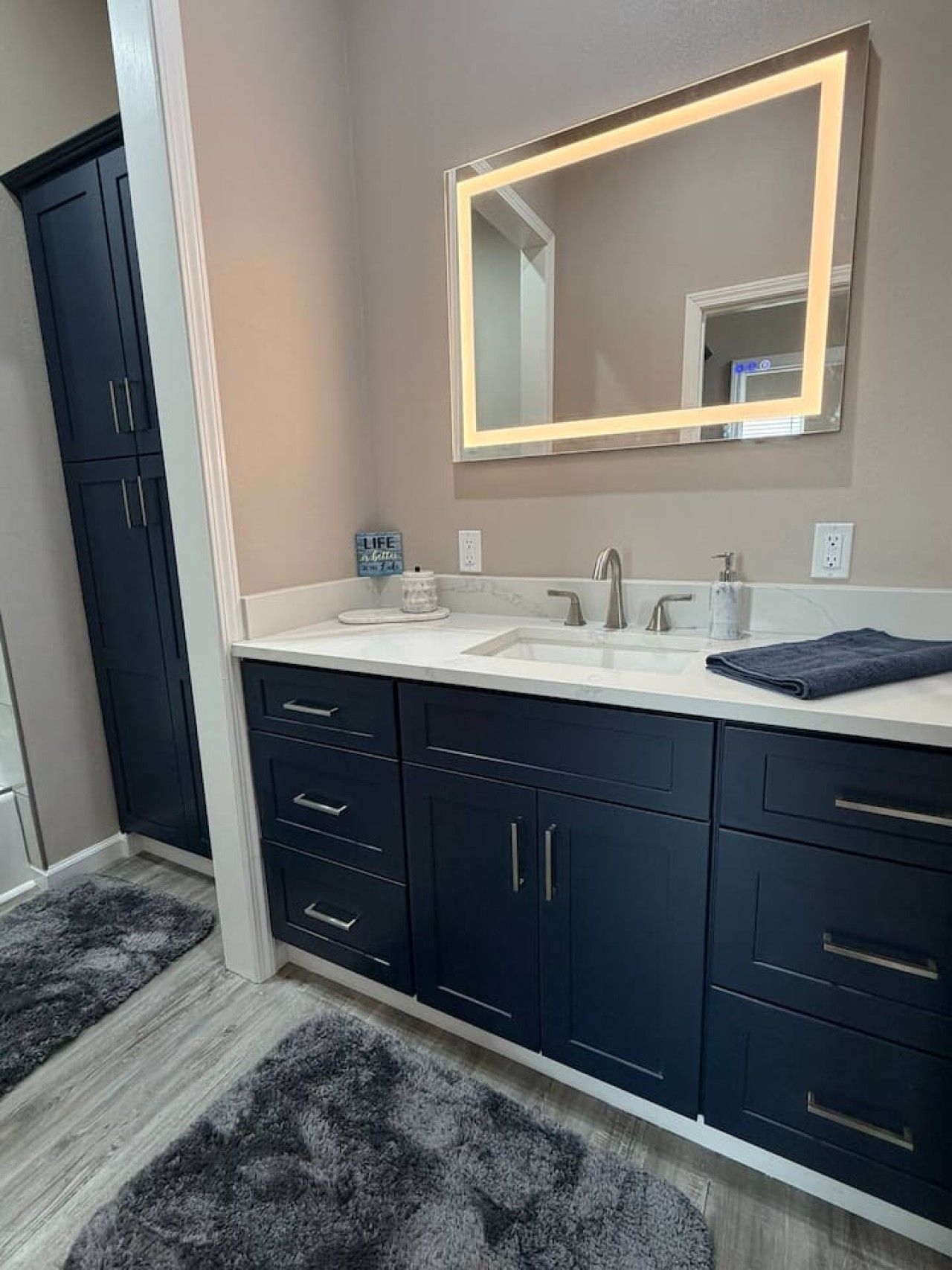 A bathroom with a double sink, dark cabinets, a mirror with a light frame, and a rug on the floor.