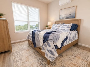 A bedroom with a bed, a nightstand, a lamp, a window with blinds, a painting, and a view of the outdoors. - Waterfront Retreat | Hot Tub, Outdoor Kitchen & Private Dock (Nocona)