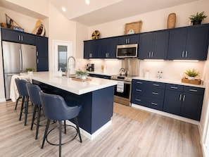 A modern kitchen with dark cabinetry, a central island with bar stools, stainless steel appliances, and decorative items on the countertops. - Waterfront Retreat | Hot Tub, Outdoor Kitchen & Private Dock (Nocona)