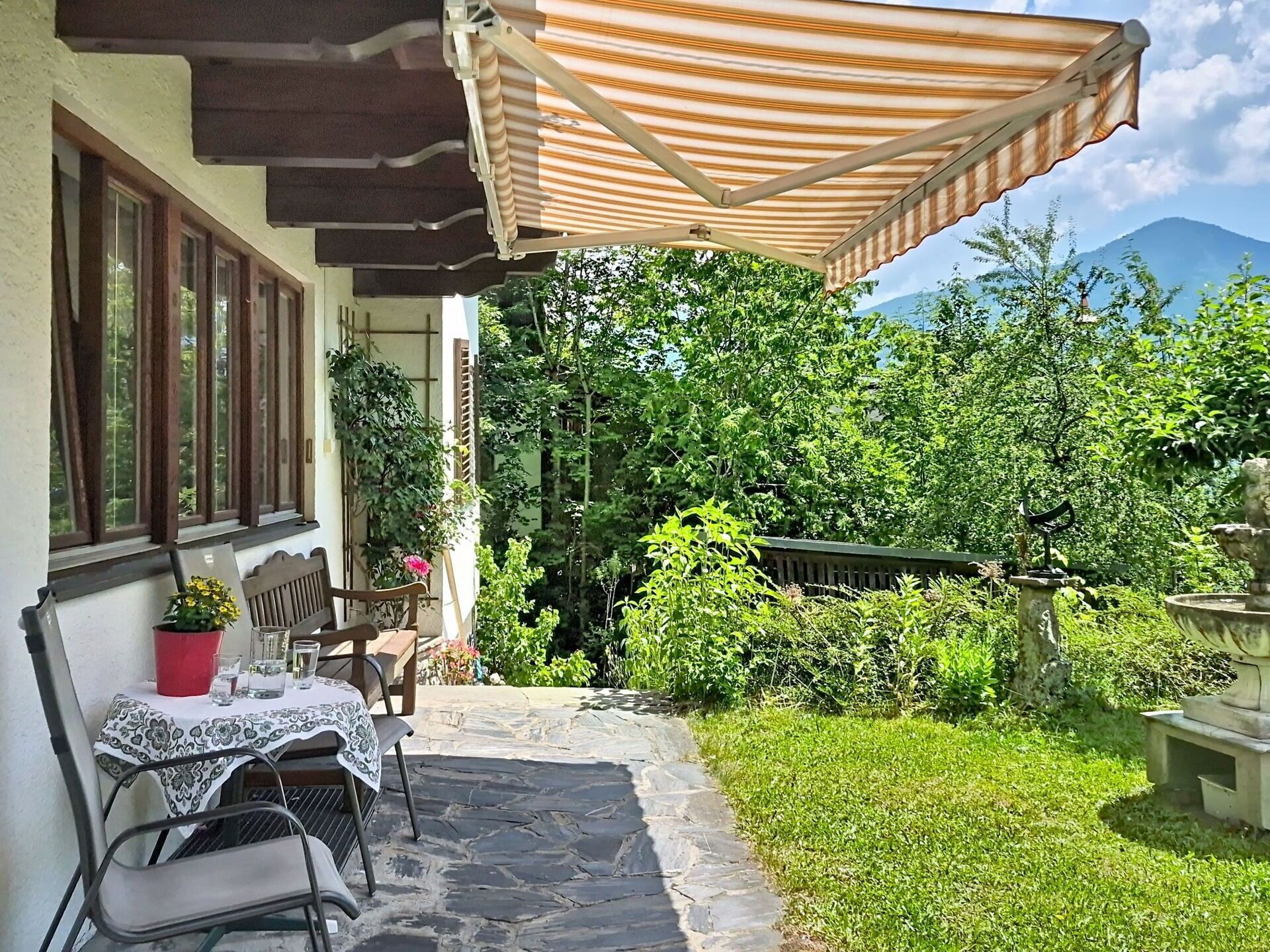 A patio with a table and chairs, a potted plant, a stone pathway, and a view of greenery and mountains.