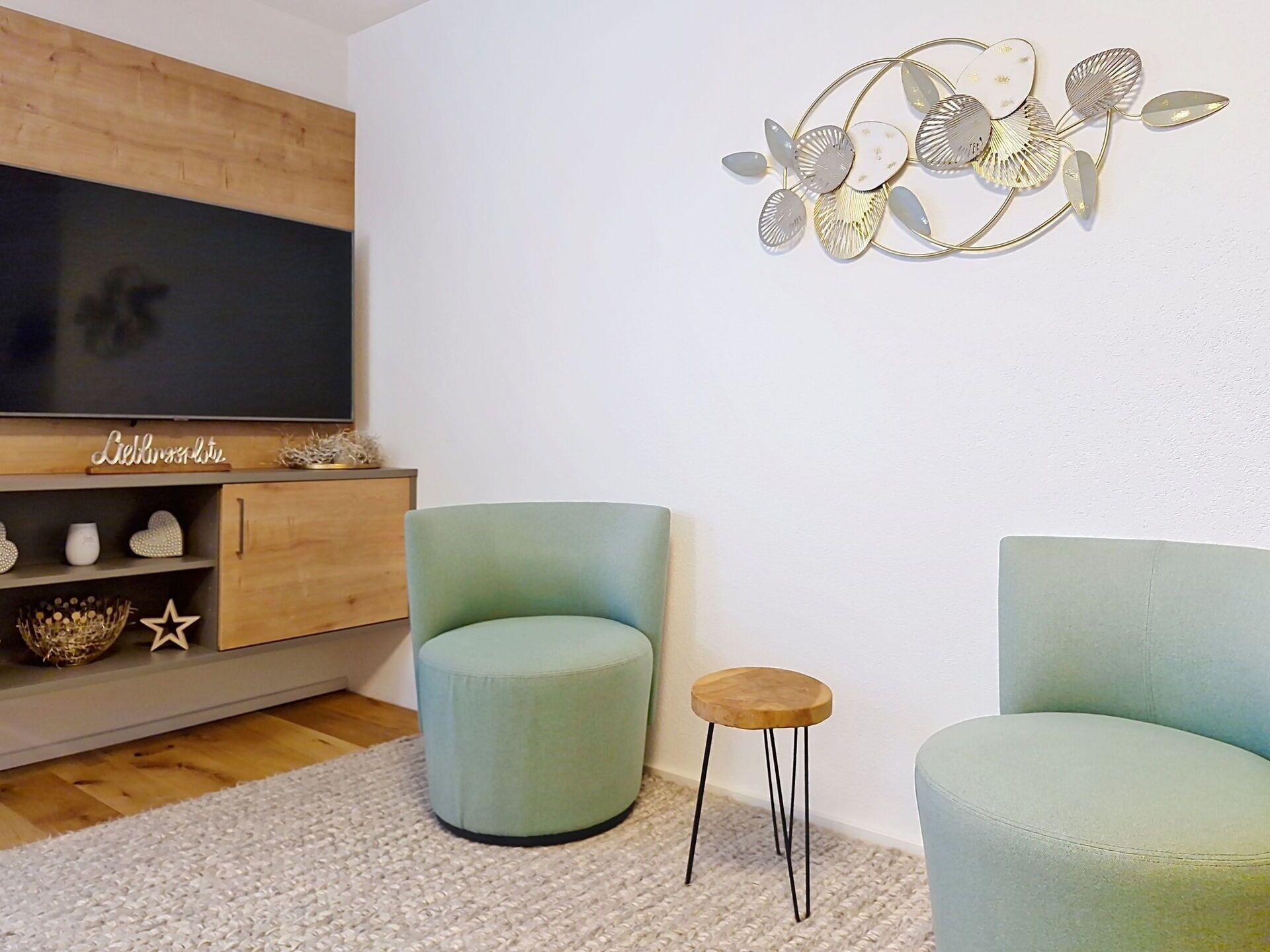 A living room with a TV, two green armchairs, a small wooden stool, and a decorative wall piece.