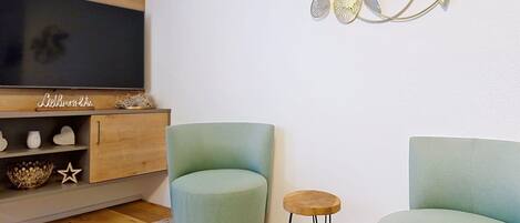 A living room with a TV, two green armchairs, a small wooden stool, and a decorative wall piece.