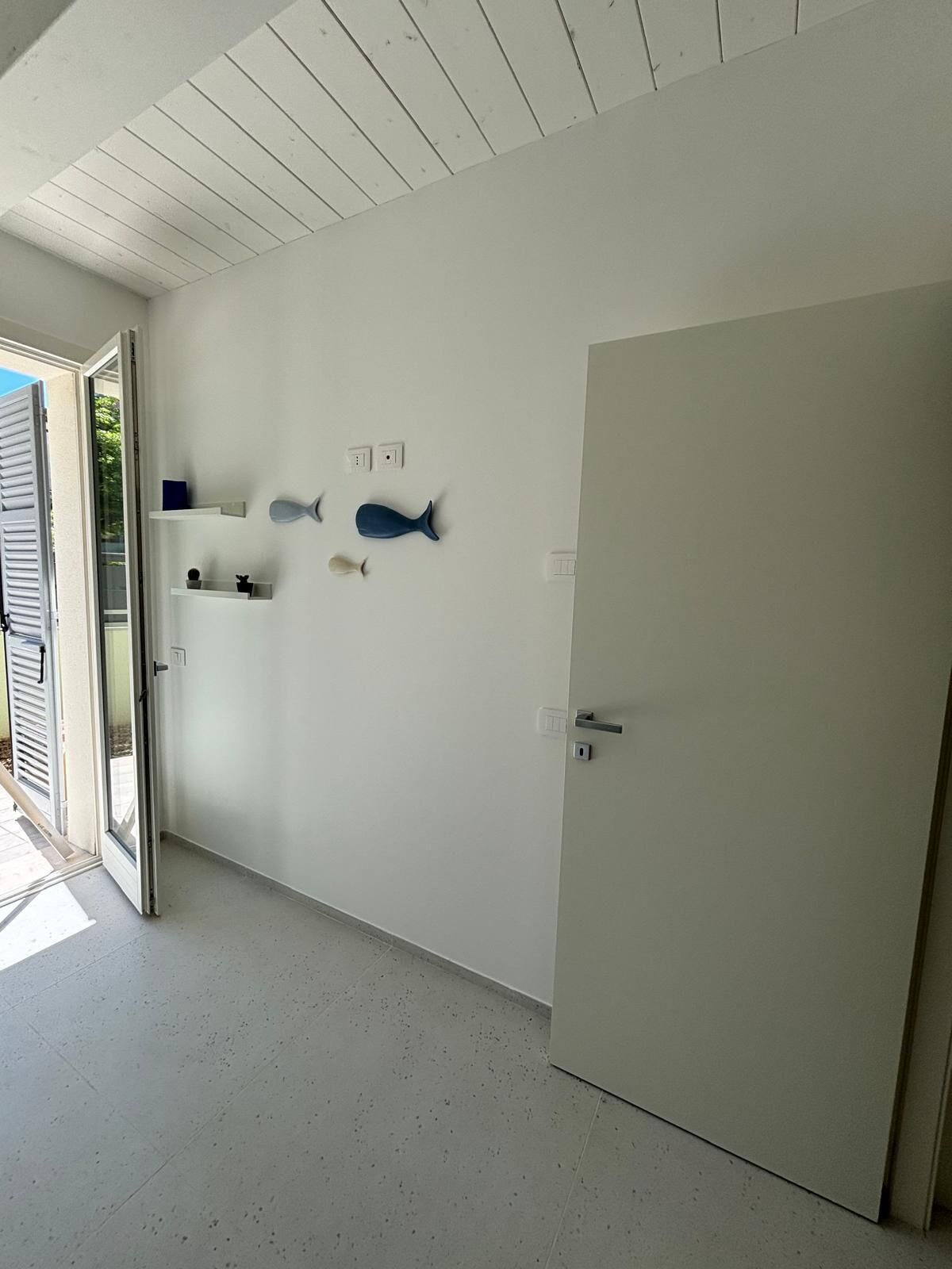 Una stanza con una porta, un soffitto e un pavimento bianchi. Sono presenti due interruttori a parete e un piccolo ripiano con un oggetto blu.