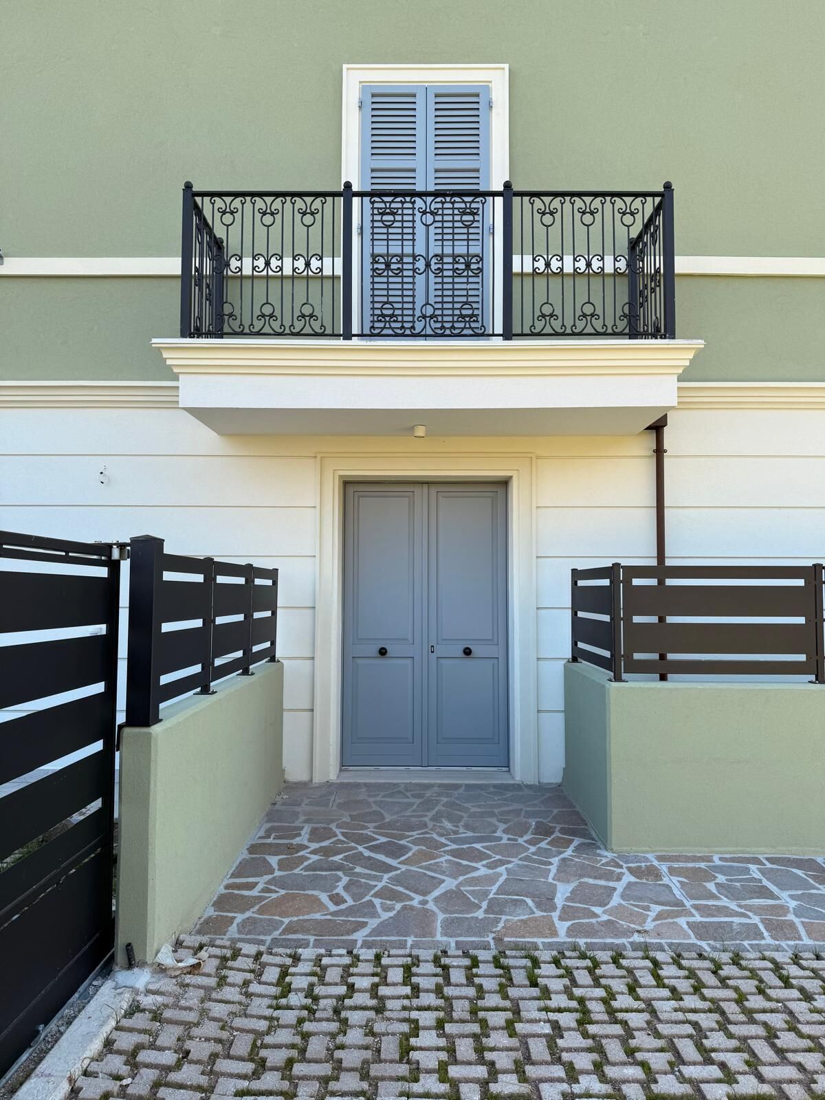 Un edificio con una porta grigia, un balcone con ringhiere nere e un ingresso con lastricato in pietra.