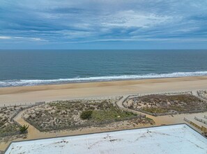 Blick auf einen Strand mit sandigem Ufer, einem eingezäunten Bereich mit Vegetation und blauem Himmel.