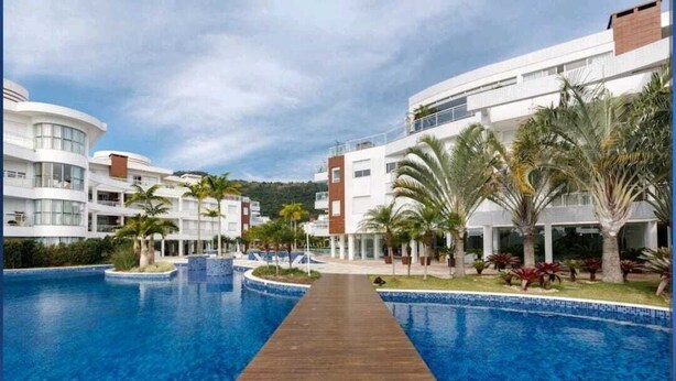 A resort with a central swimming pool, connected by a wooden walkway, surrounded by palm trees and modern buildings. - Gustavo Funchal (Florianópolis)