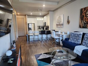 A modern living room with a glass coffee table, a blue sofa, and a kitchen area with white cabinets and a stainless steel refrigerator. - De Premium Place Buckhead- City Center (Atlanta)