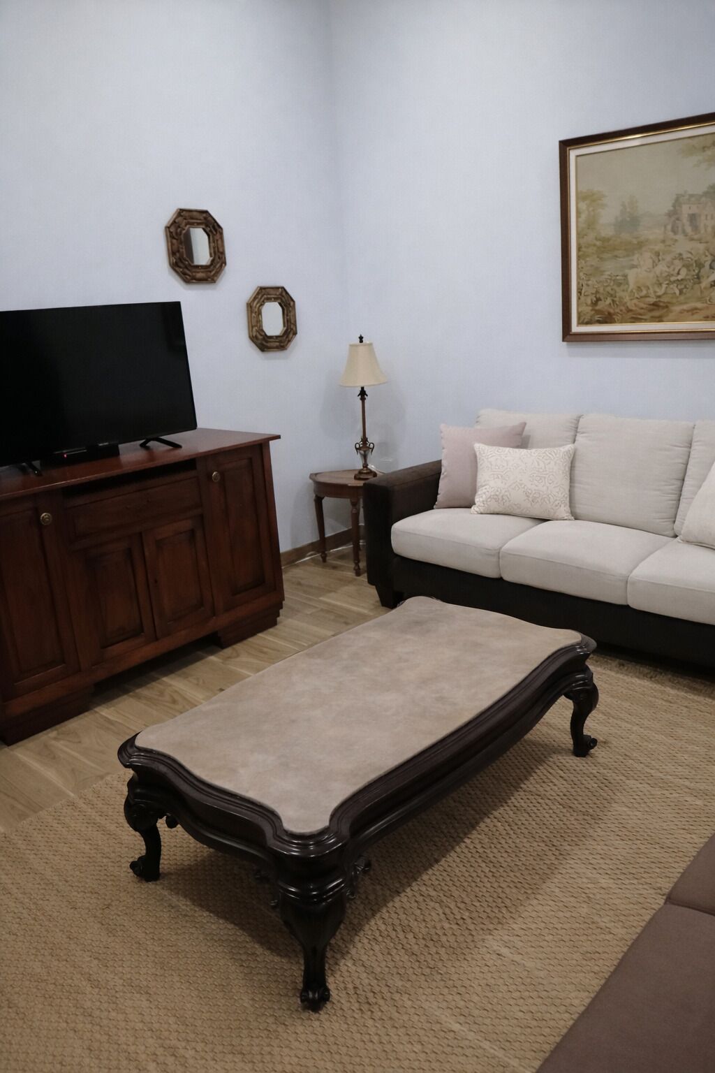 Een woonkamer met een televisie op een houten kast, een beige bank, een klein bijzettafeltje met een lamp en een ingelijst schilderij aan de muur.