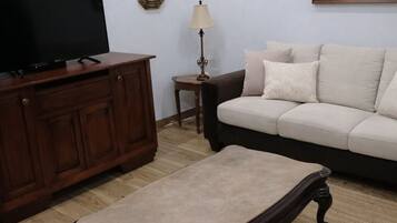 Een woonkamer met een televisie op een houten kast, een beige bank, een klein bijzettafeltje met een lamp en een ingelijst schilderij aan de muur.