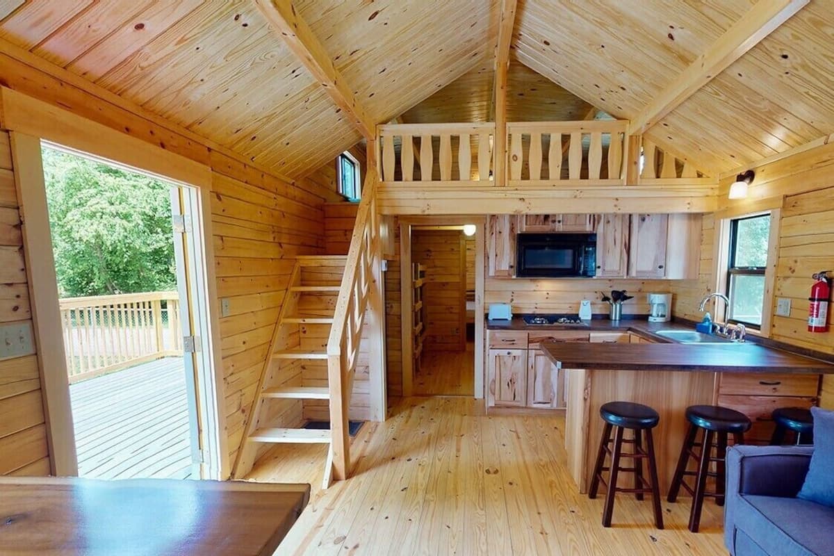 A wooden cabin interior with a kitchen, dining area, and a loft bedroom.