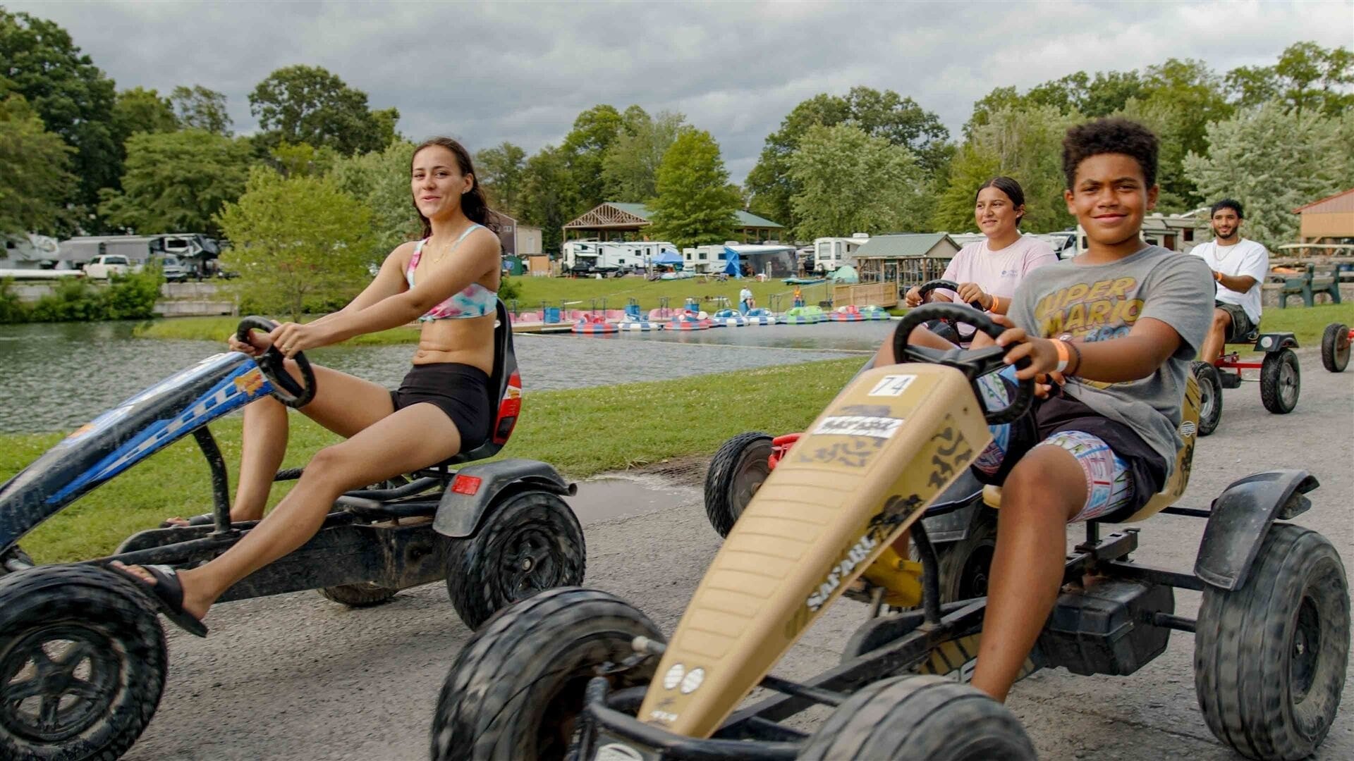 Personas conduciendo karts sobre un camino pavimentado cerca de una masa de agua, con vehículos recreativos y árboles al fondo.