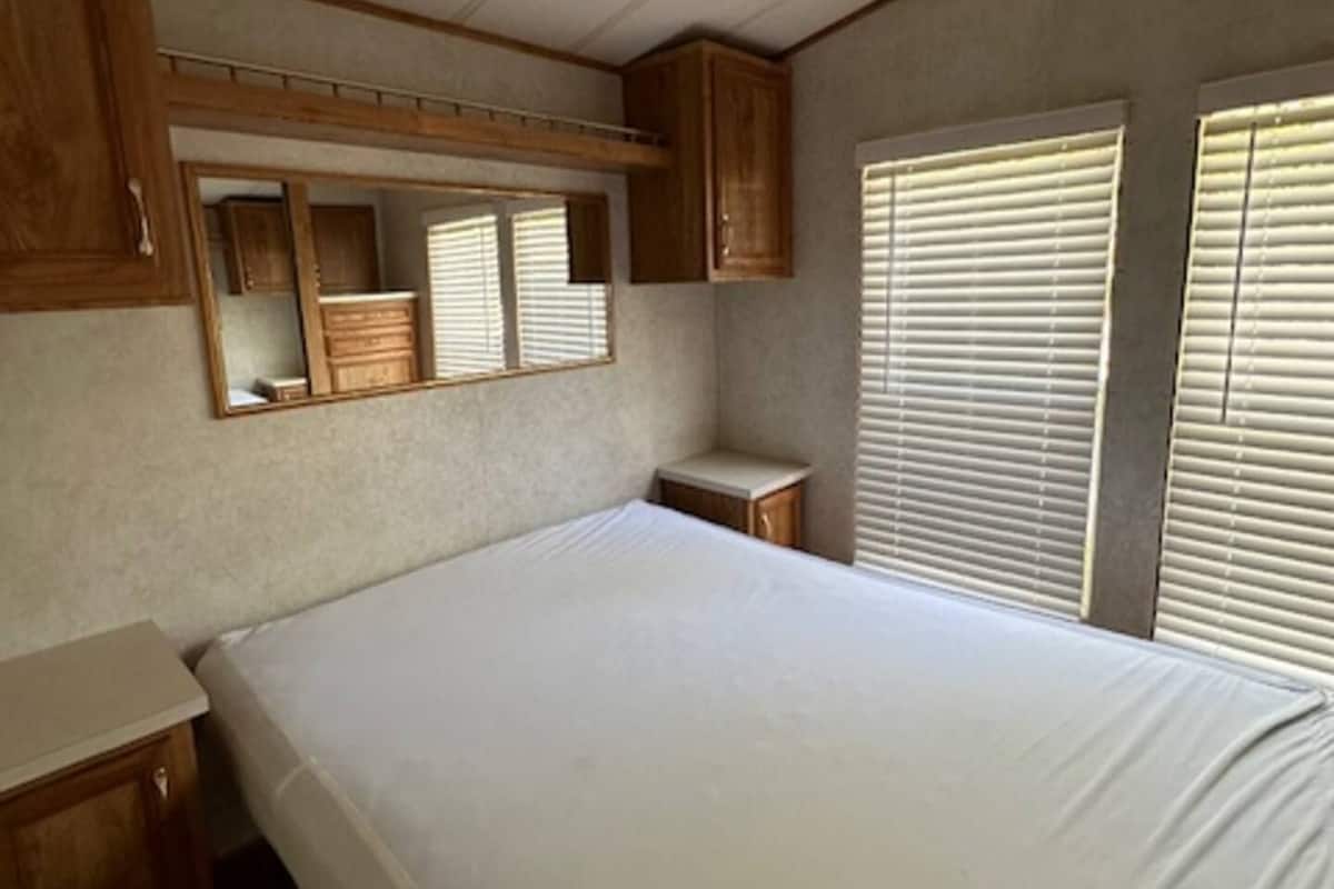 A bedroom with a single bed, wooden headboard, a mirror, and two windows with blinds.