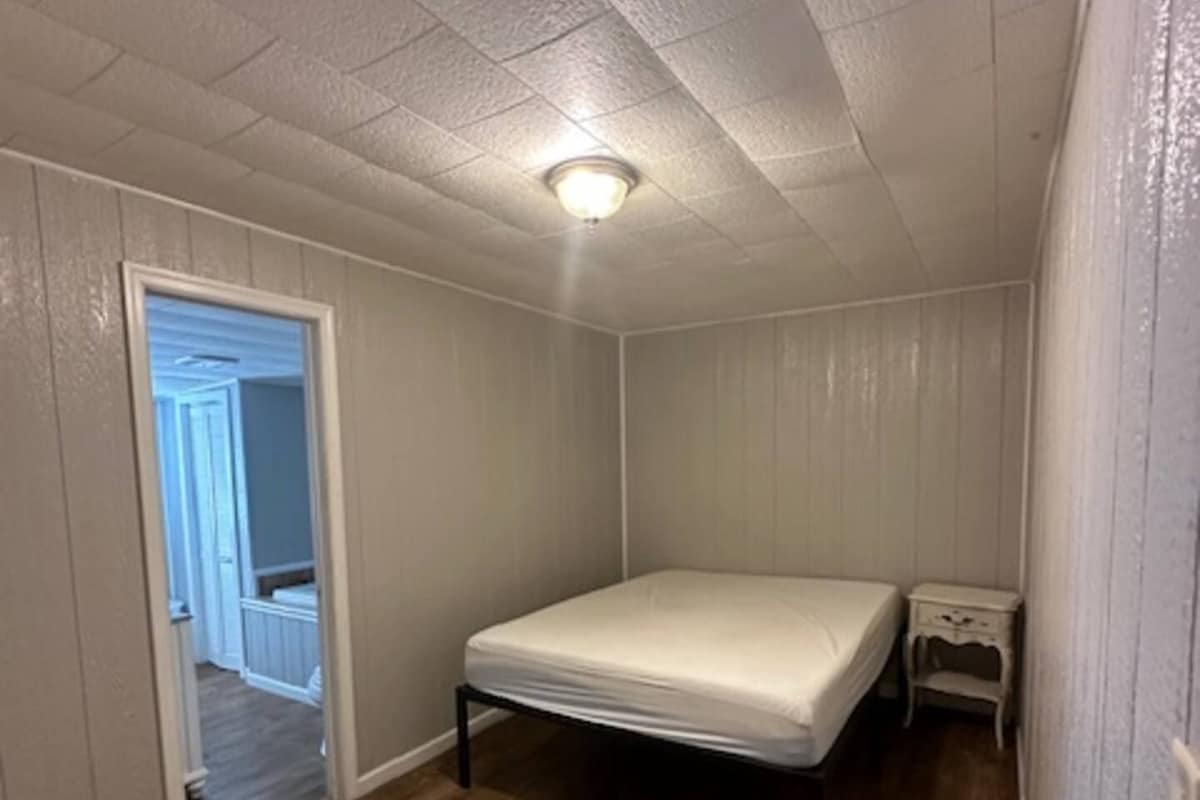 A small, single-bed room with a wooden floor, a white ceiling, and a door leading to another room.