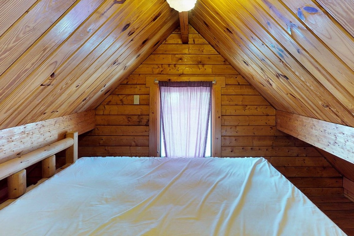A wooden attic bedroom with a bed, a window with curtains, and a sloped ceiling.