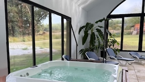 A hot tub in a room with large windows, a wooden bench, and a view of the outdoors.