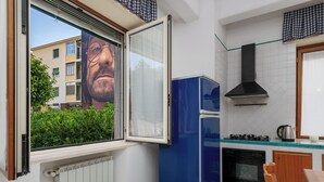Een kleine keuken met een blauwe koelkast, een gasfornuis en een raam met uitzicht op een gebouw en groen.