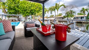 A covered outdoor seating area with wicker furniture, a red tray with a champagne bottle and glasses, and a view of a marina with boats and palm trees. - Luxury Fort Lauderdale Waterfront Home • Heated Pool • Game Room •  Park (Fort Lauderdale)