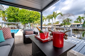 A covered outdoor seating area with wicker furniture, a red tray with a champagne bottle and glasses, and a view of a marina with boats and palm trees. - Luxury Fort Lauderdale Waterfront Home • Heated Pool • Game Room •  Park (Fort Lauderdale)
