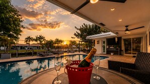 A poolside area with a bottle of champagne in a red bucket, two wine glasses, and a view of a sunset over a swimming pool. - Luxury Fort Lauderdale Waterfront Home • Heated Pool • Game Room •  Park (Fort Lauderdale)