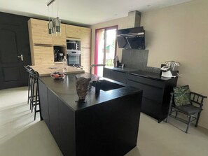 A modern kitchen with a central island, built-in appliances, and a dining area with a chair. - Le Minihic sur Rance - Charming house with its cat Platon between land and sea (Le Minihic-sur-Rance)