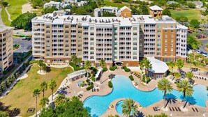 A multi-story hotel with a pool area, including a large pool and a smaller pool, surrounded by palm trees and a golf course in the background. - FREE Daily Golf! Reflections 406 Amazing Views, Heated Resort Pool & Hot Tub! (Panama City)