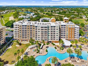 Ein mehrstöckiges Hotel mit Poolbereich, inklusive eines großen und eines kleineren Pools, umgeben von Palmen und einem Golfplatz im Hintergrund.