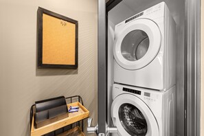 A laundry area with stacked washing machines, a framed corkboard on the wall, and a wooden tray with items. - FREE Daily Golf! Reflections 406 Amazing Views, Heated Resort Pool & Hot Tub! (Panama City)