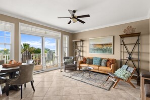 A spacious living room with a large window, a ceiling fan, a leather sofa, and a dining area with chairs and a table. - FREE Daily Golf! Reflections 406 Amazing Views, Heated Resort Pool & Hot Tub! (Panama City)