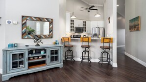 Una cocina moderna con armarios blancos, un microondas y un refrigerador. Hay dos taburetes de bar con asientos de madera y estructura metálica. Se ve un ventilador de techo, y lámparas colgantes sobre la isla de la cocina.