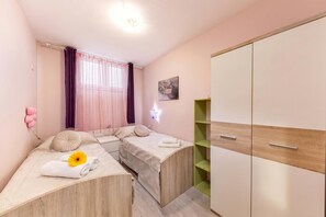 A bedroom with two beds, a wardrobe, a window with curtains, and a wall-mounted light. - Pool and Garden Apartment Hortenzija (Kostrena)