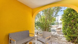 A room with a yellow wall, a grey sofa, a round table with a plate of food, and a view of greenery through an archway. - Pool and Garden Apartment Hortenzija (Kostrena)