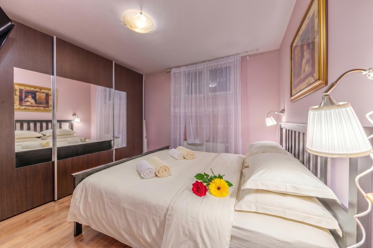 Een hotelkamer met een bed, een spiegel, een bedlampje en een raam met dunne gordijnen.