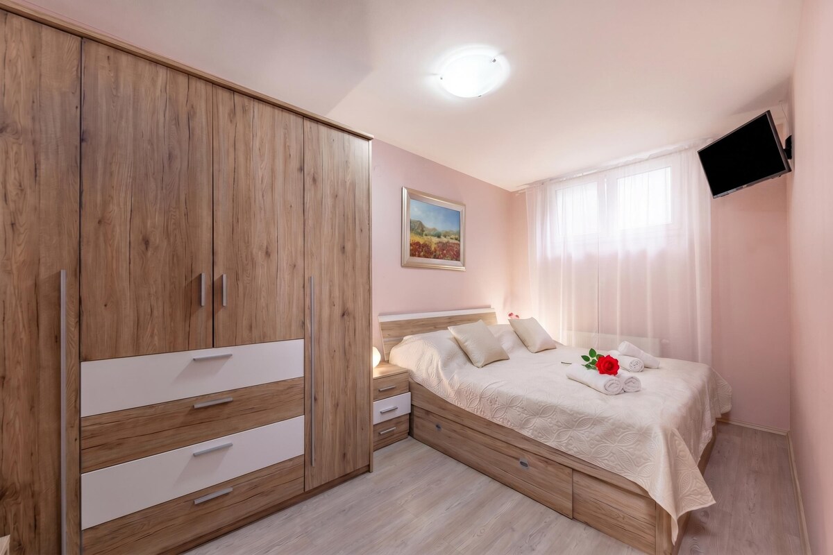 Een slaapkamer met een houten garderobe, een bed met een witte dekbedovertrek, een nachtkastje, een schilderij aan de muur, en een aan de muur bevestigde televisie.