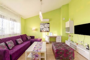 A living room with a purple sofa, a white coffee table, a desk, and a television. - Pool and Garden Apartment Hortenzija (Kostrena)