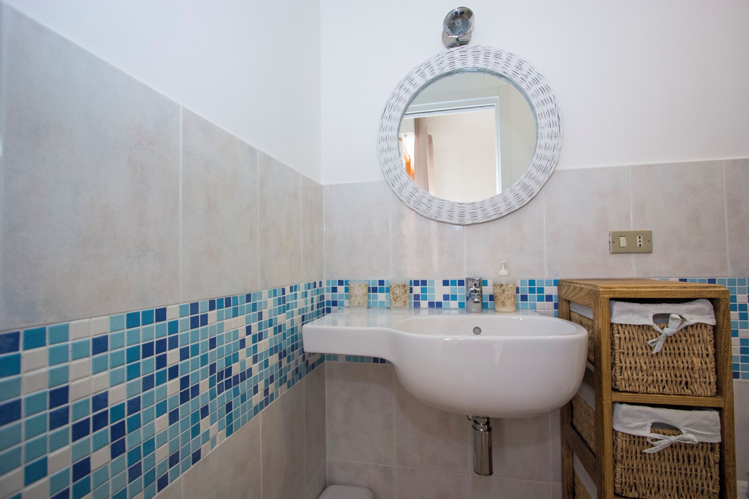 Ein Badezimmer mit einem weißen Waschbecken, einem runden Spiegel und einer gefliesten Wand mit blau und beige farbenen Fliesen.