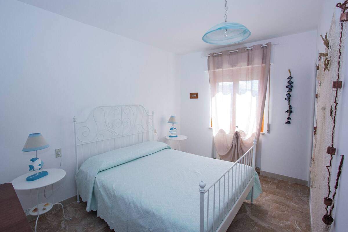 A bedroom with a bed, bedside tables, a lamp, a window with curtains, and a wall with decorative elements.