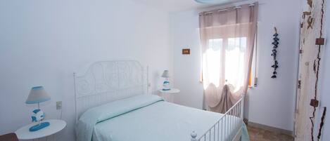 Un dormitorio con cama, mesitas de noche, una lámpara, una ventana con cortinas y una pared decorada.